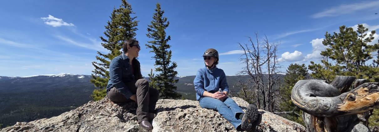 Two women sitting on a rocky mountain overlook surrounded by pine trees and snow-capped peaks during a guided trail ride near Dubois,