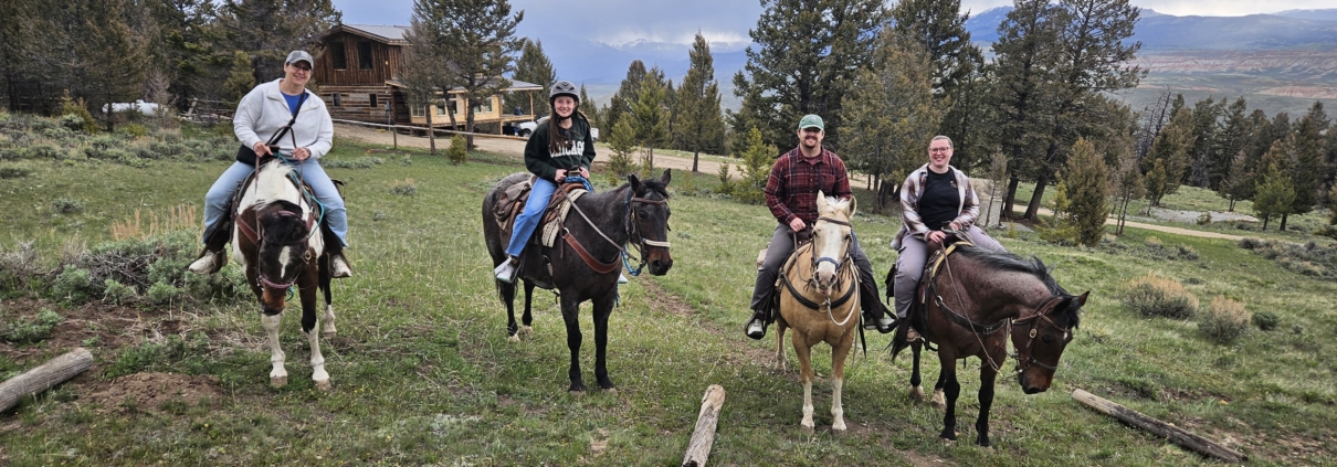 Four guests on horseback in front of a log cabin with mountain and canyon views at a Wyoming guest ranch