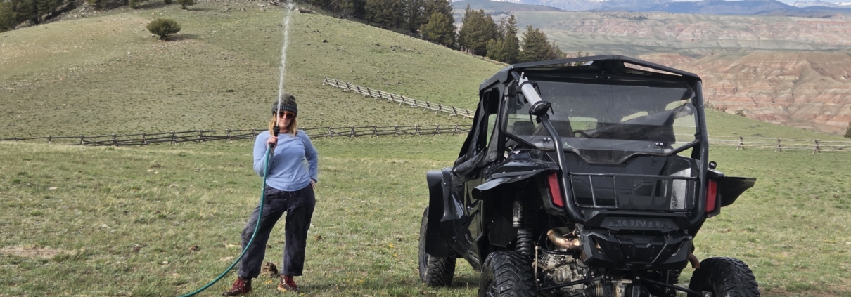 Ranch staff washing a muddy side-by-side UTV on a green hillside with canyon and mountain views near Dubois, Wyoming