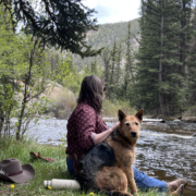 Guide sitting in the grass and early spring flowers in the canyon beside a river with a red and black working dog, cowboy hat and water bottle nearby