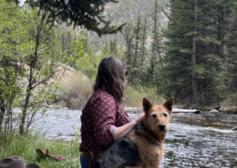 Guide sitting in the grass and early spring flowers in the canyon beside a river with a red and black working dog, cowboy hat and water bottle nearby