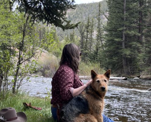 Guide sitting in the grass and early spring flowers in the canyon beside a river with a red and black working dog, cowboy hat and water bottle nearby