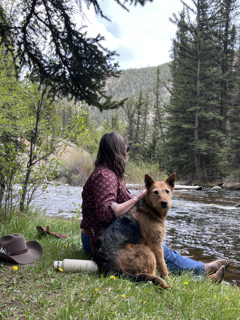 Guide sitting in the grass and early spring flowers in the canyon beside a river with a red and black working dog, cowboy hat and water bottle nearby